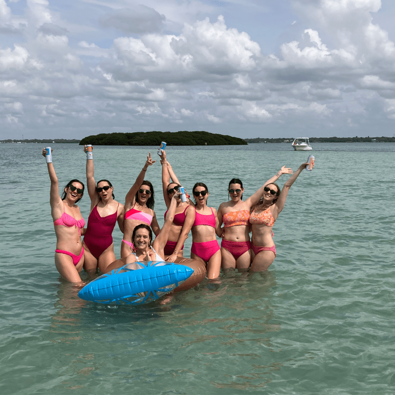 Seven people in swimsuits smiling in shallow water, holding drinks, with clouds and an island in the background.