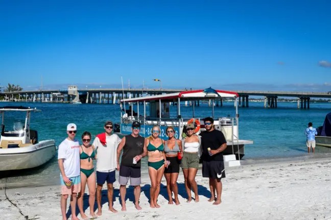Group of people in swimwear posing on a sandy beach with boats and bridge in background.